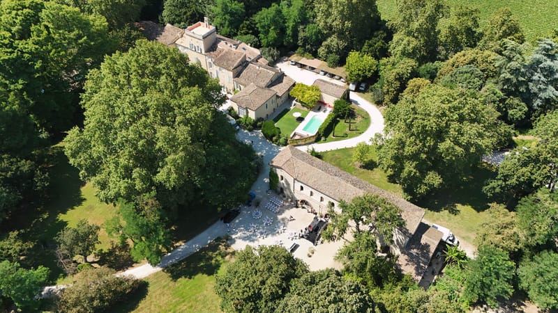 An aerial drone shot of a French country estate being set up for a wedding reception. The property features a multi-building stone complex with terracotta-tiled roofs, a rectangular swimming pool surrounded by manicured hedging, and a curved driveway with parked vehicles. In the lower courtyard area adjacent to a long stone barn with arched openings, numerous white-draped round tables and chairs are arranged in preparation for an outdoor reception, with several people visible moving around the setup. A vineyard is visible in the upper right corner of the frame. Potential venue feature image.