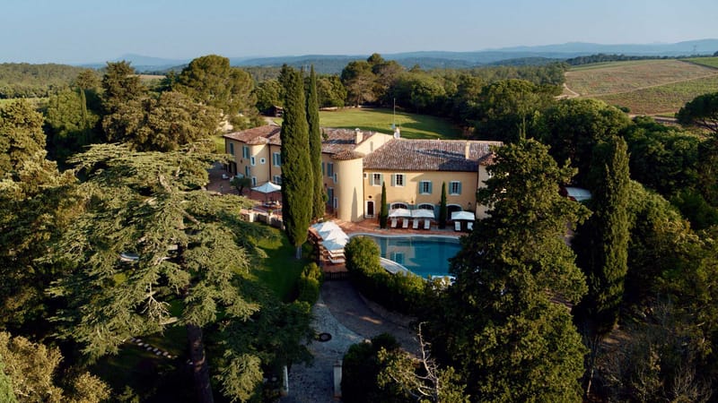 An aerial wide shot of a large Provençal bastide-style property with warm yellow ochre rendered walls, terracotta tile roofs, teal-painted shutters, and a distinctive round tower. The grounds include a large rectangular outdoor pool with white sun loungers and parasols arranged around the terrace, a gravel driveway leading to the entrance, and formal landscaping with tall Italian cypress trees flanking the main façade. The property is surrounded by mature woodland and opens onto open lawns toward the rear, with vineyards visible on the hillside to the right. No people are present in the image. Potential venue feature image.