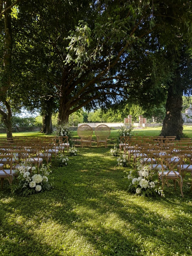 Outdoor ceremony set beneath mature trees, with wooden chairs and white florals