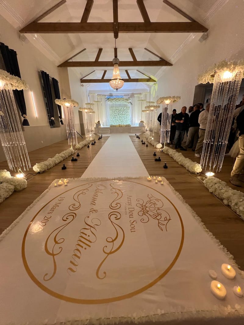 An indoor wedding ceremony aisle set with a white monogrammed runner, crystal strand decorations and candles, under a beamed ceiling.