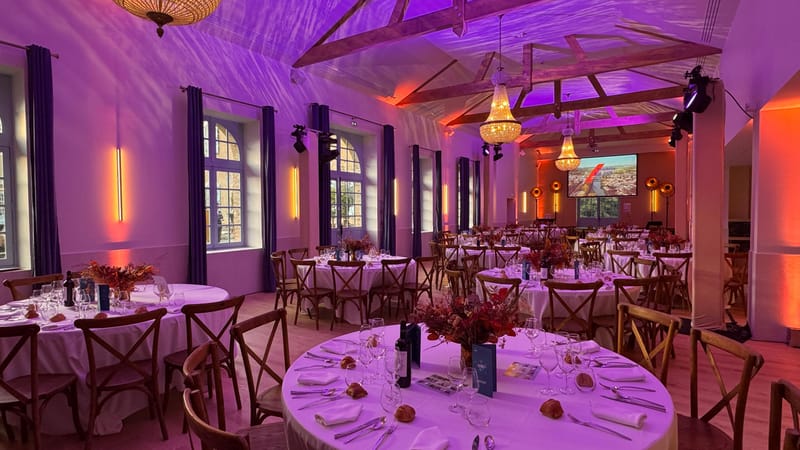 The Orangerie set for a formal dinner with round tables and floral centrepieces, lit in purple and orange under a beamed ceiling.