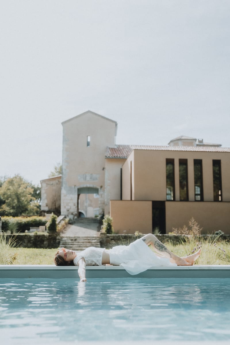 A bride in a white dress reclining at the edge of the swimming pool, with the château's modern extension building behind.