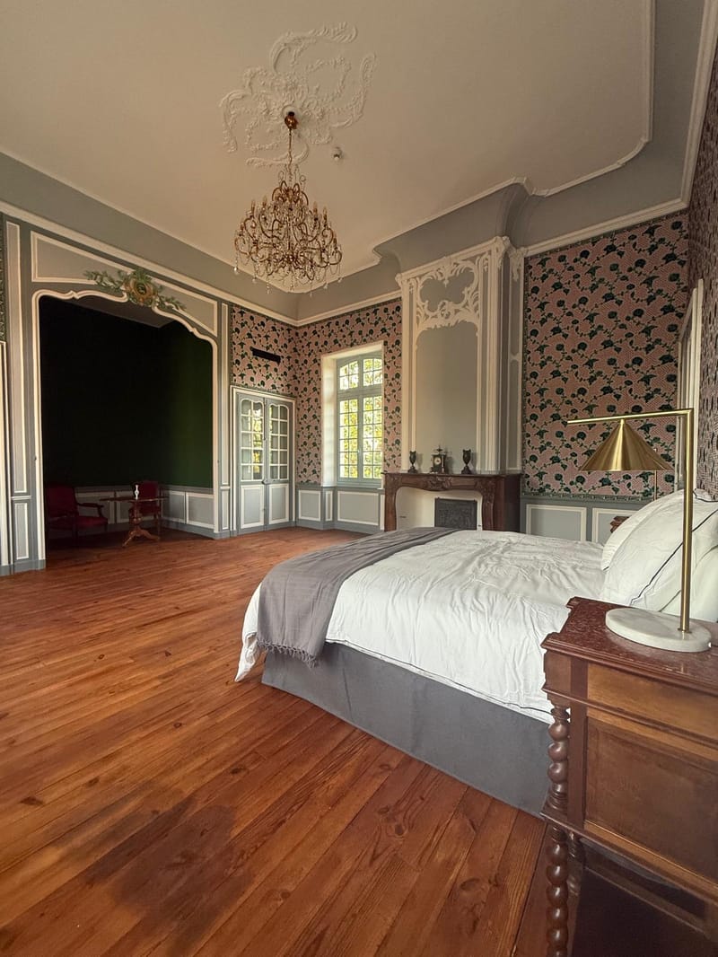 A bedroom with floral wallpaper, a crystal chandelier and a large bed with white linens.