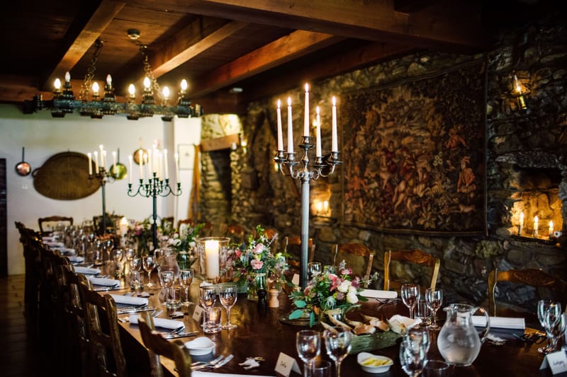 Long table set for dinner in the candlelit dining room, with a stone wall and tapestry