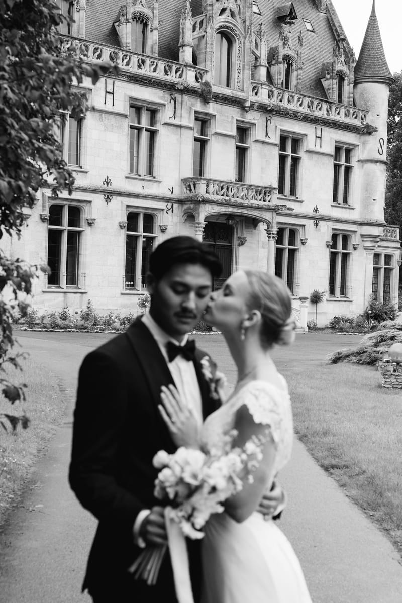 A bride kissing the groom on the cheek, bouquet in hand, in front of the château facade, in black and white