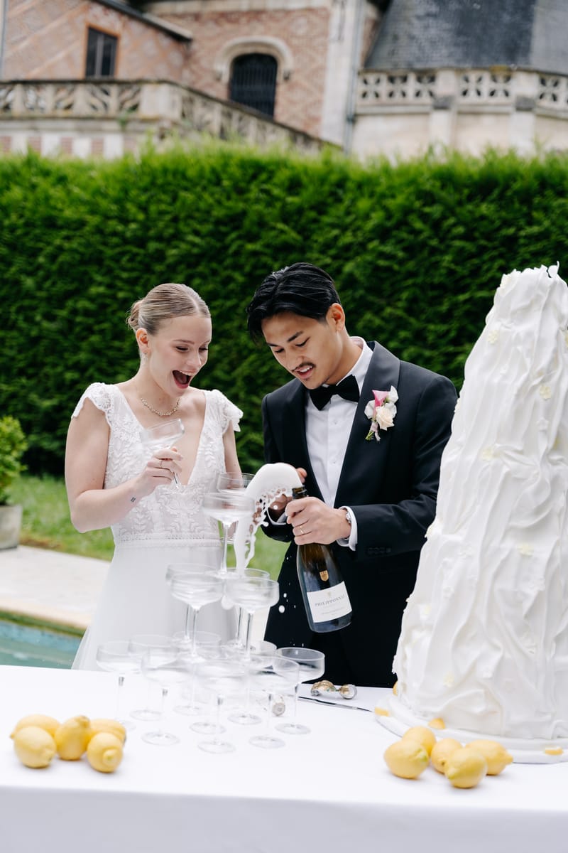 A bride laughing, coupe in hand, as the groom pours champagne over a coupe tower by the pool, beside a tall white cake