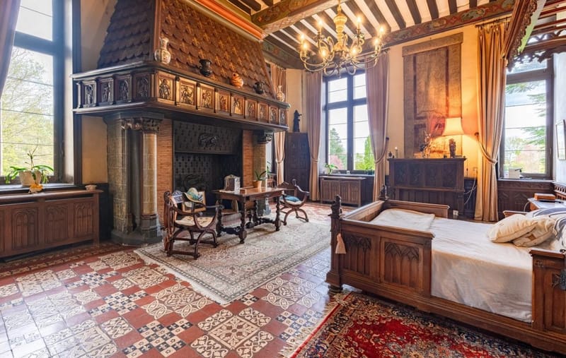 The Louis XII bedroom with a carved oak bed, painted beamed ceiling, hooded fireplace and patterned tile floor