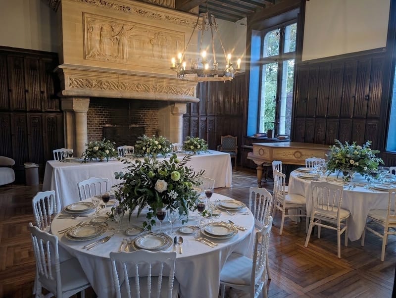 Round tables set for dinner in the Pulligny salon, under a candle chandelier beside the stone fireplace and piano