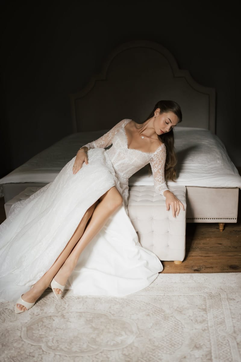 A bride in a lace gown seated on a tufted bench at the foot of a bed