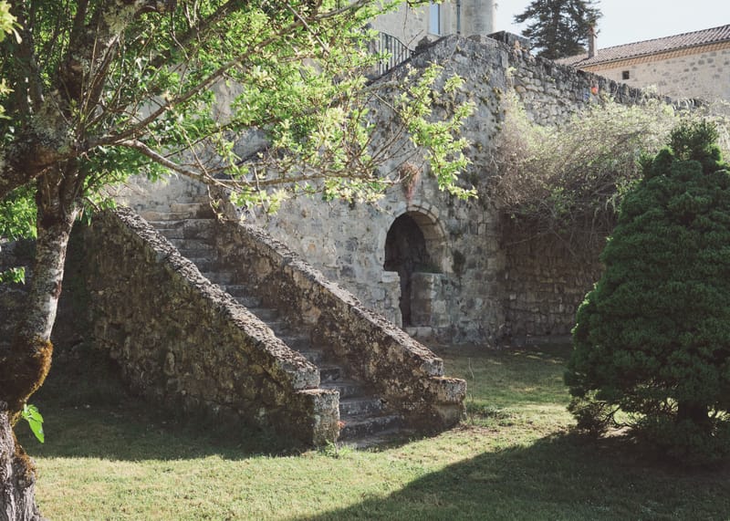 Stone staircase between mossy parapet walls, climbing beside an arched doorway under the branches of a tree