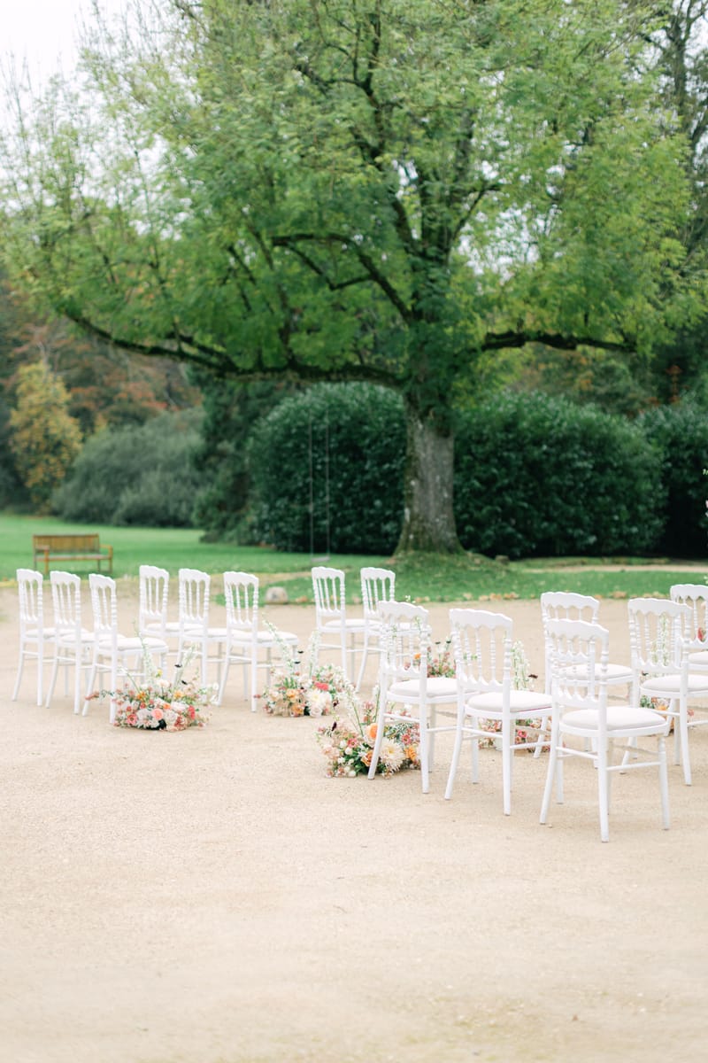 Outdoor ceremony setup with Napoleon chairs and floral aisle markers under a large tree at Château de Méridon