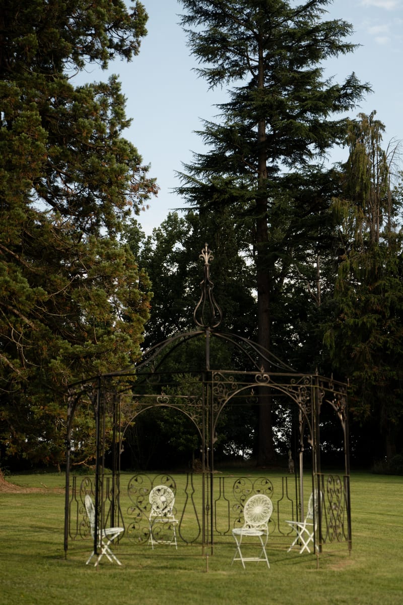 Wrought-iron gazebo with chairs in the gardens of Château de Méridon, framed by tall trees