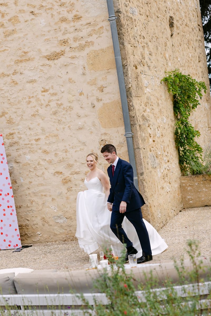 Bride and groom walk hand in hand across gravel at the foot of a tall honey-coloured stone wall