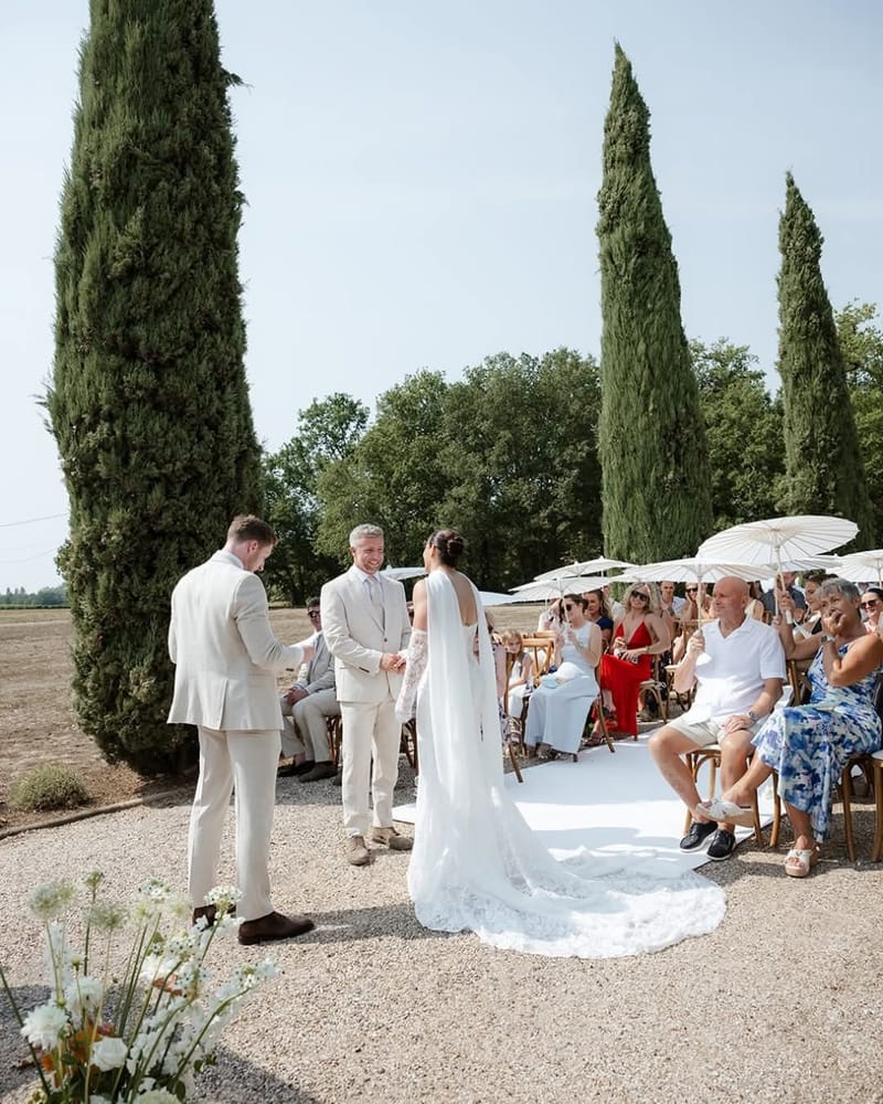 Bride faces a smiling man in a cream suit, a second man in cream beside them, guests with parasols seated by cypresses