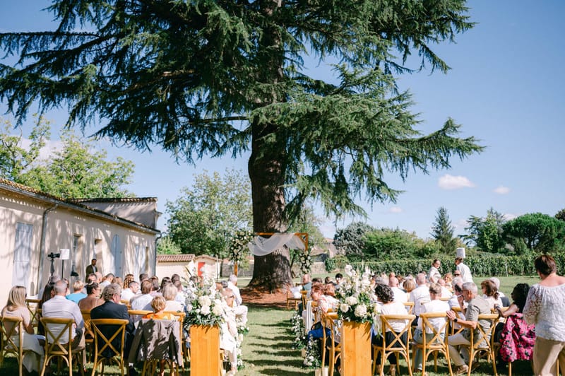 Outdoor ceremony setup under a mature cedar tree with seated guests in the walled park at Chateau de l'Hospital