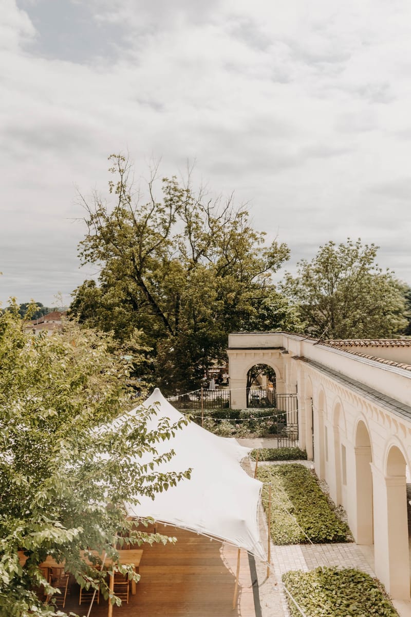 Stretch tent reception set up against the arcades at Chateau de l'Hospital with long wooden tables and festoon lighting