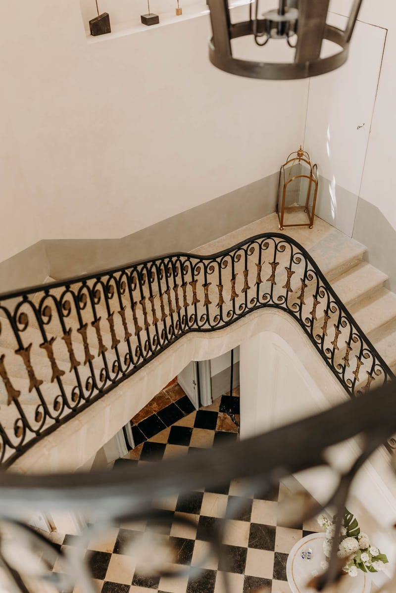 Top-down view of the wrought-iron grand staircase at Chateau de l'Hospital with a gold pendant lantern and chequerboard marble floor