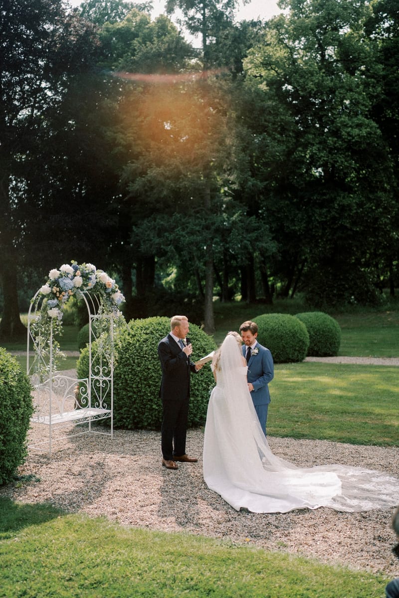 A ceremony at the white metal arbour with the officiant and guests among the topiary