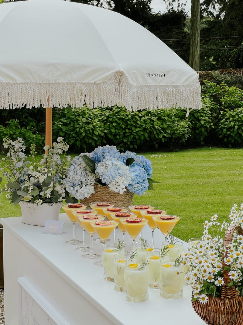 A drinks table set with cocktails and hydrangeas under a fringed parasol on the lawn