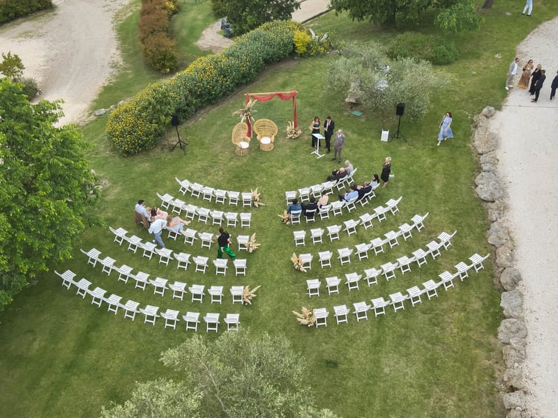 Aerial view of a lawn set with curved rows of white folding chairs facing a draped arch, with two wicker chairs and a lectern beside a flowering hedge.