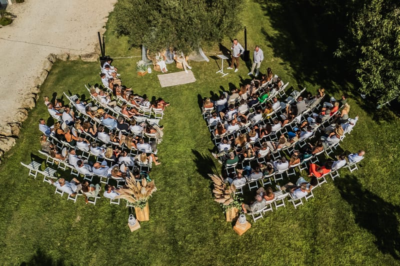 Aerial view of a ceremony on the lawn, guests seated in two blocks of white chairs facing a draped arch beneath olive trees.