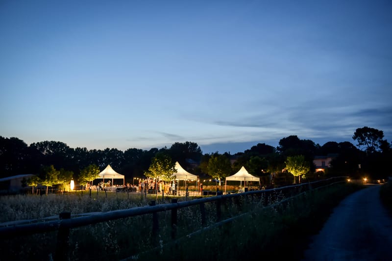 White event marquees glowing with lights on the lawn at dusk, seen across a field.