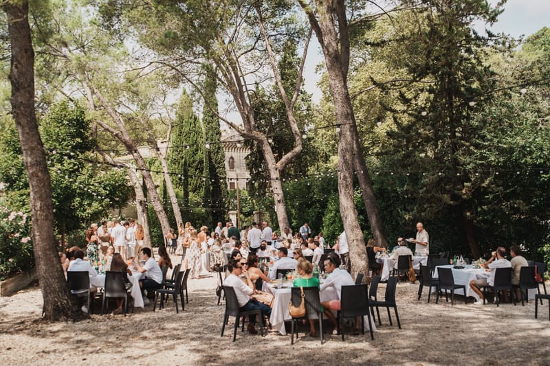 Guests dining at white-clothed tables under tall pine trees strung with festoon lights, a stone building beyond.