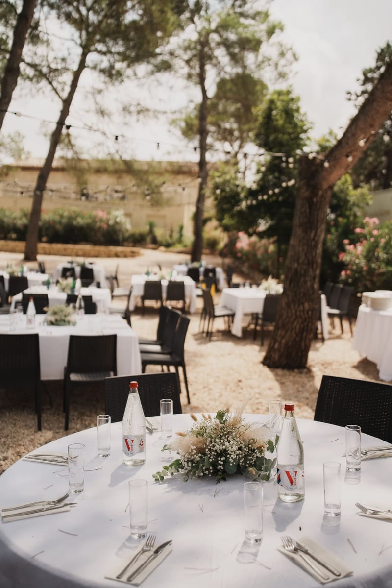 Round table set with white linen, glassware and a posy of gypsophila and pampas, with further set tables on gravel beneath pine trees.