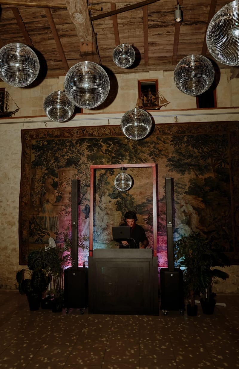 A DJ booth set up in front of a large tapestry beneath a beamed ceiling hung with mirrored disco balls