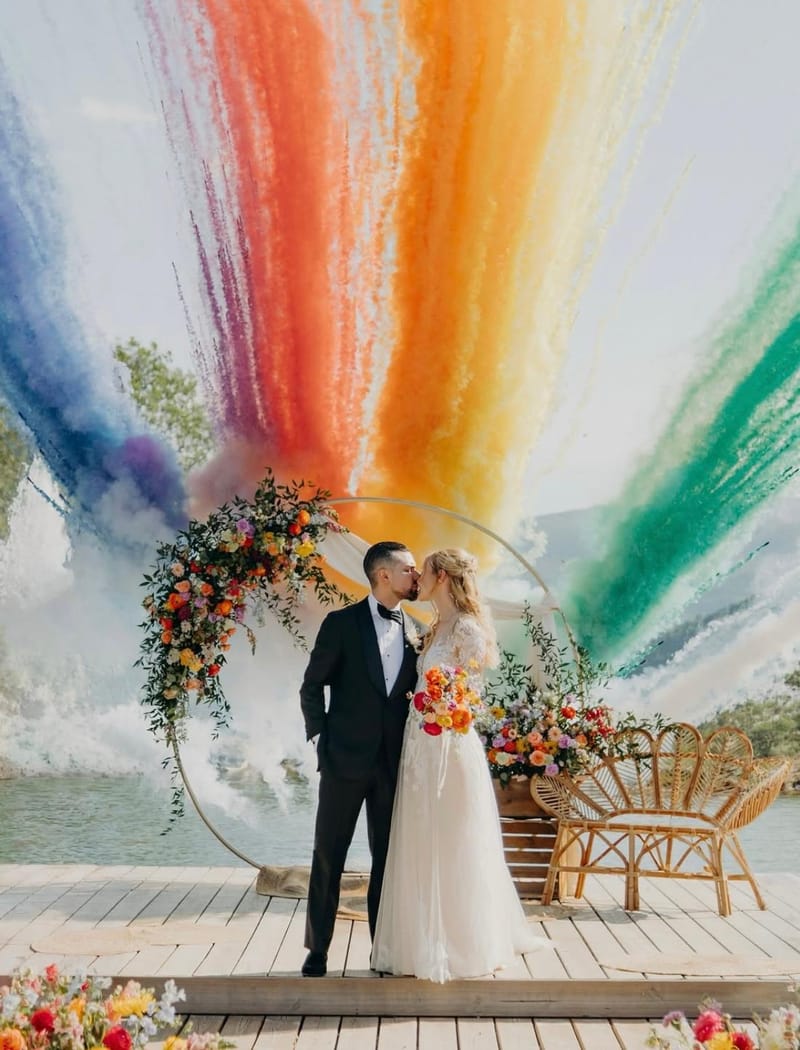 A couple kiss in front of a circular floral arch on the lakeside deck at Le Mas des Cinq Fontaines as coloured smoke rises behind them.