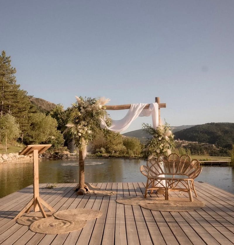 Wooden ceremony arch dressed with dried florals and white drapery on the lakeside deck at Le Mas des Cinq Fontaines at golden hour.