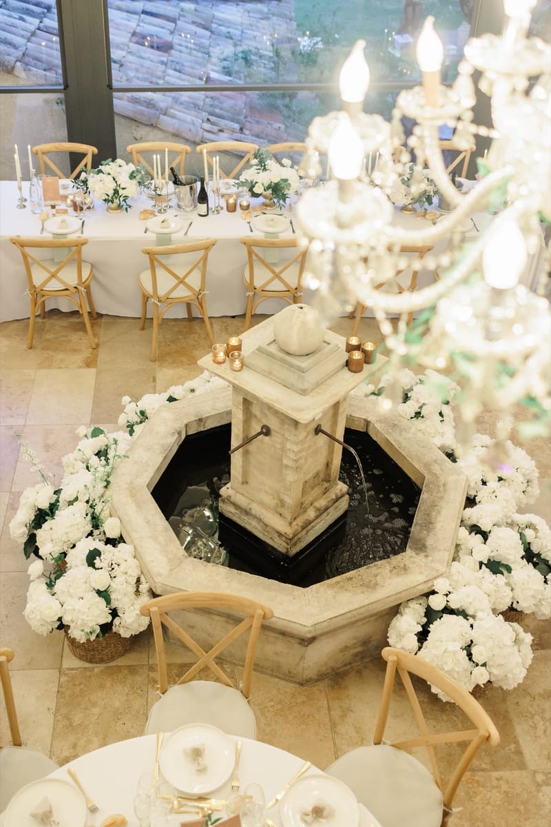 Wedding tables set around the stone fountain in the orangery at Le Mas des Cinq Fontaines beneath crystal chandeliers.