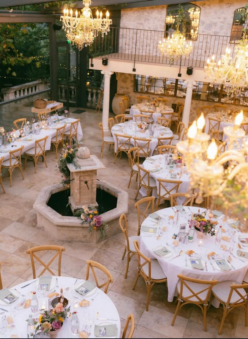 The orangery at Le Mas des Cinq Fontaines set for a wedding dinner with round tables, cross-back chairs and chandeliers.
