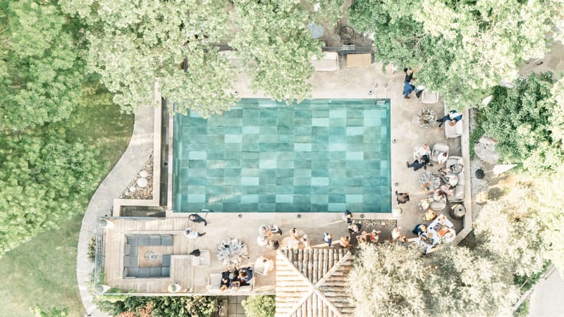 Aerial view of the swimming pool and shaded terrace at Le Mas des Cinq Fontaines with guests gathered poolside under the trees.