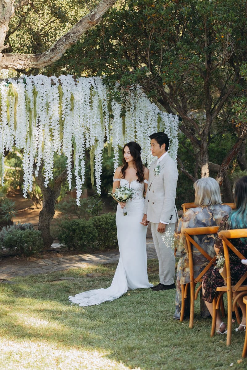 Wedding ceremony beneath the olive trees with hanging white florals at Les Oliviers de Palombaggia