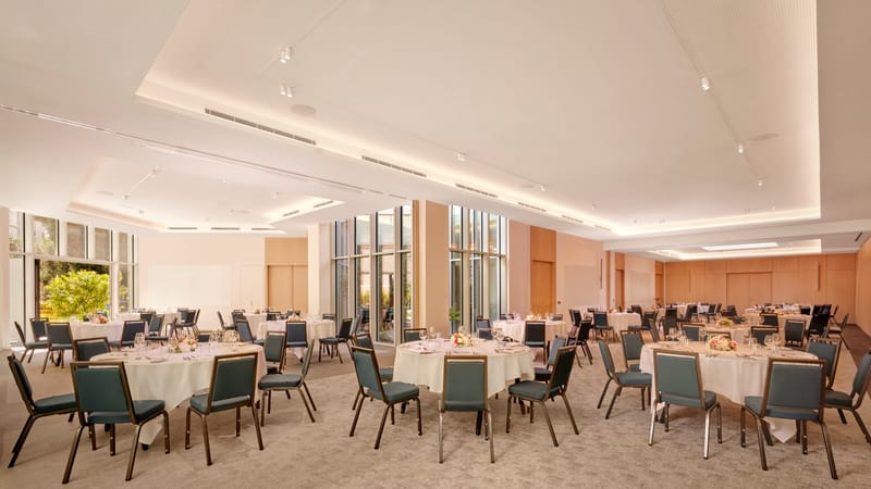 Event room set with round tables, white linen and blue-grey chairs beside tall garden windows