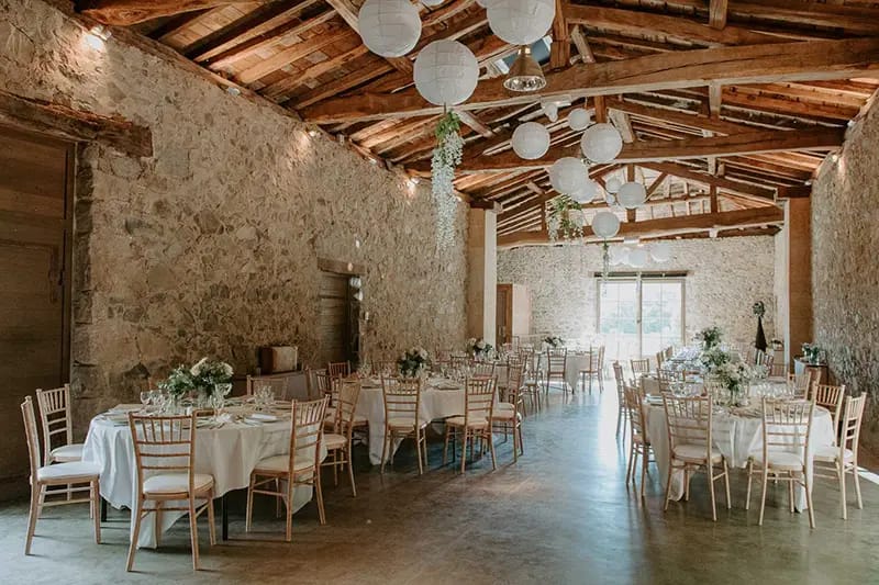 Round tables set for a wedding reception in the stone barn at Domaine de Vavril, under a beamed roof